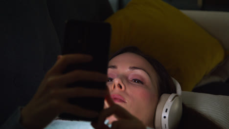 Woman-Wearing-Wireless-Headphones-Lying-On-Sofa-At-Home-At-Night-Streaming-Music-Or-Watching-Movie-On-Mobile-Phone-2